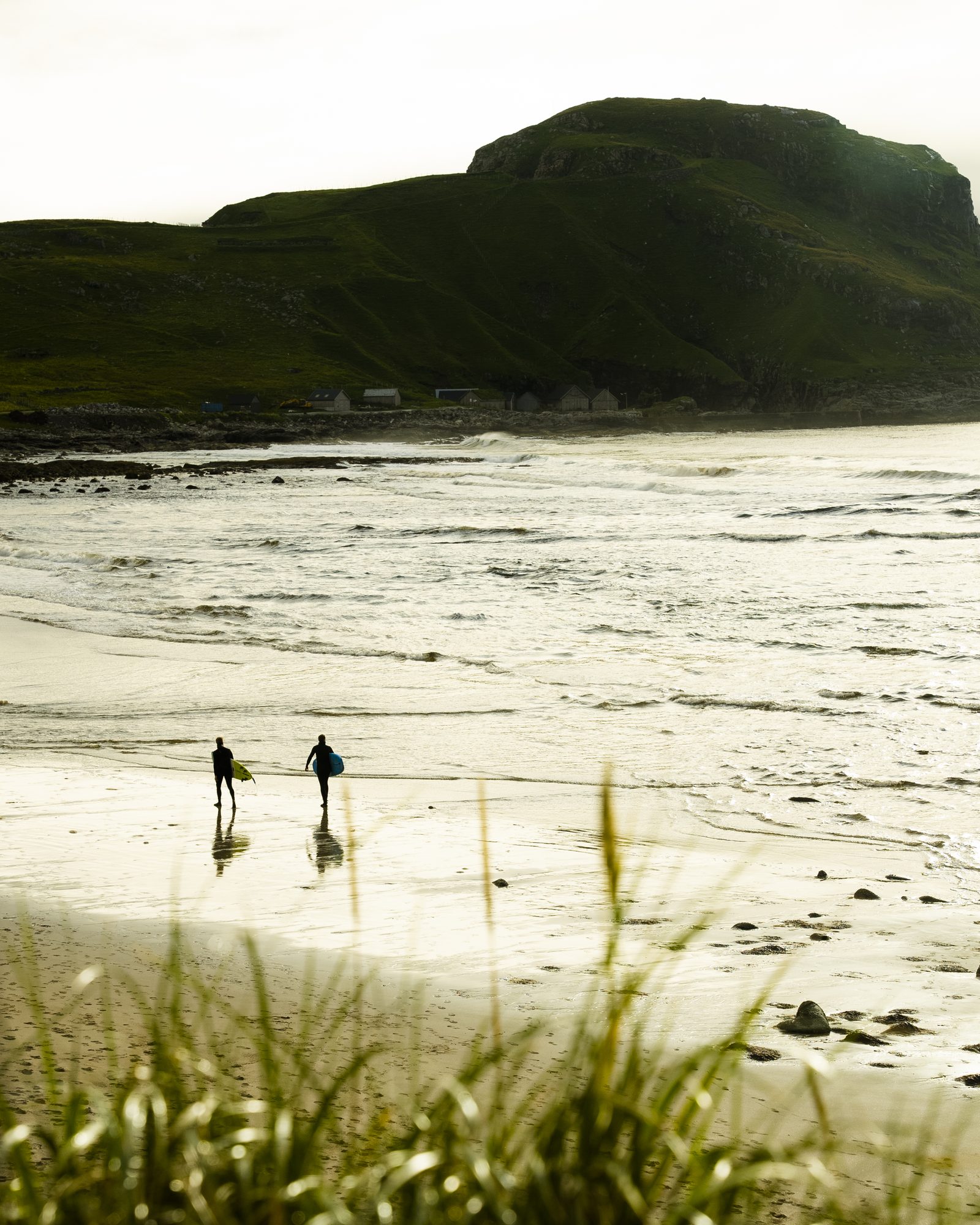 Two surfers walking a beach below a green headland