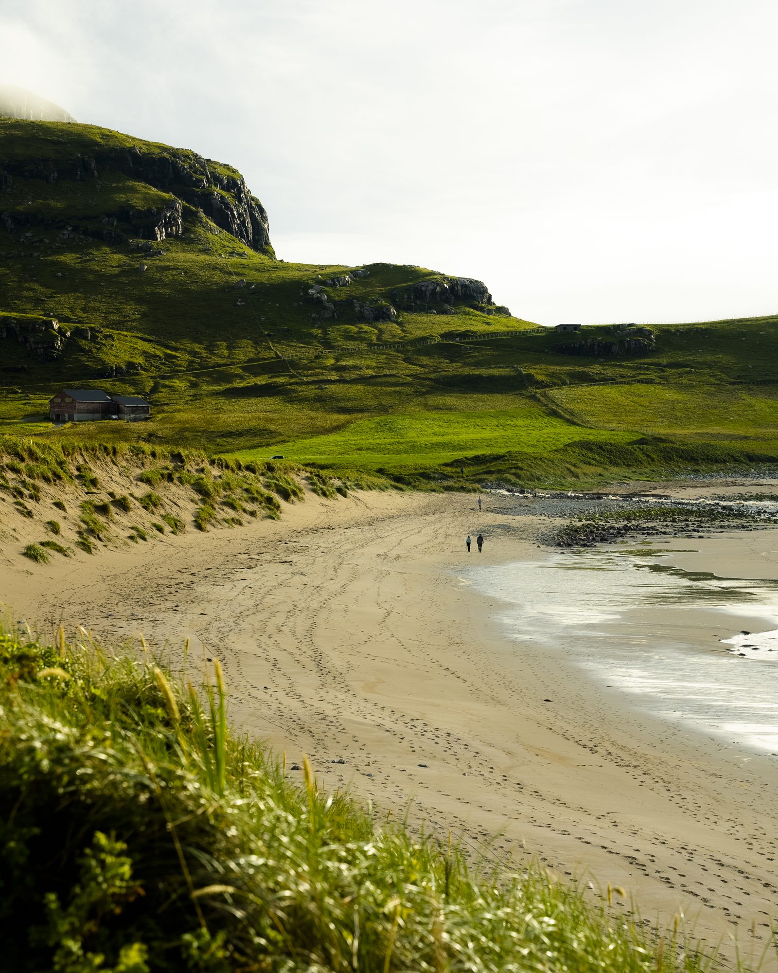 Two people walking an empty beach below green hills